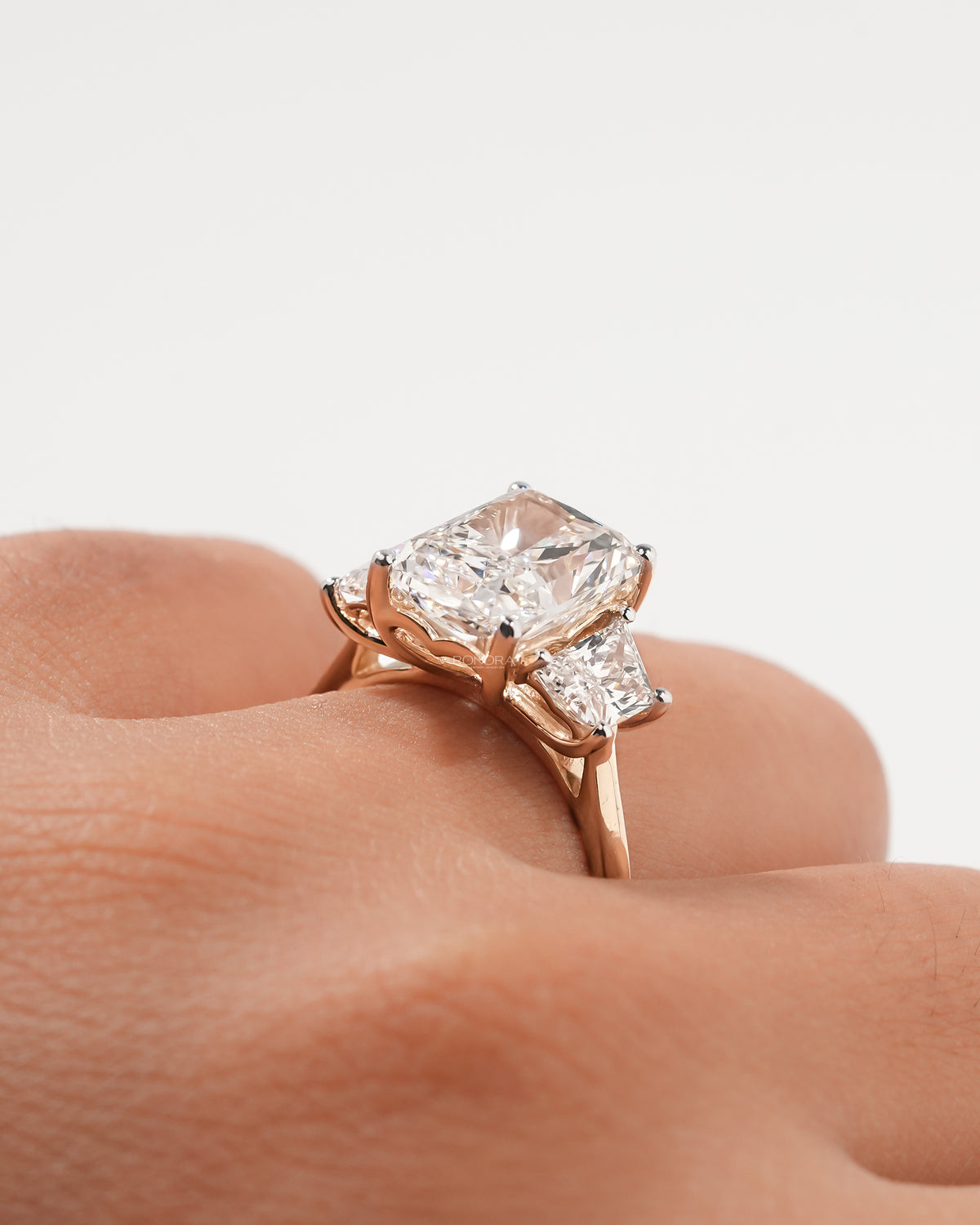 Close-up of a rose gold radiant cut lab grown diamond three stone ring with trapezoid side stones on a finger