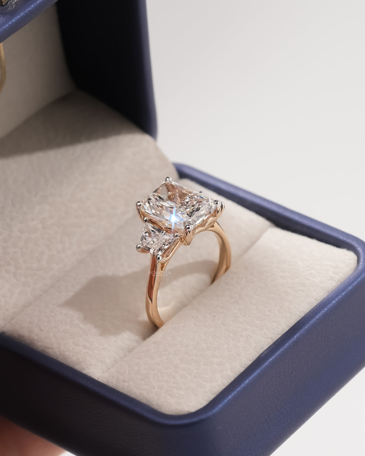 Rose gold radiant cut lab grown diamond three stone ring with sparkle, shown at an angle in a navy ring box