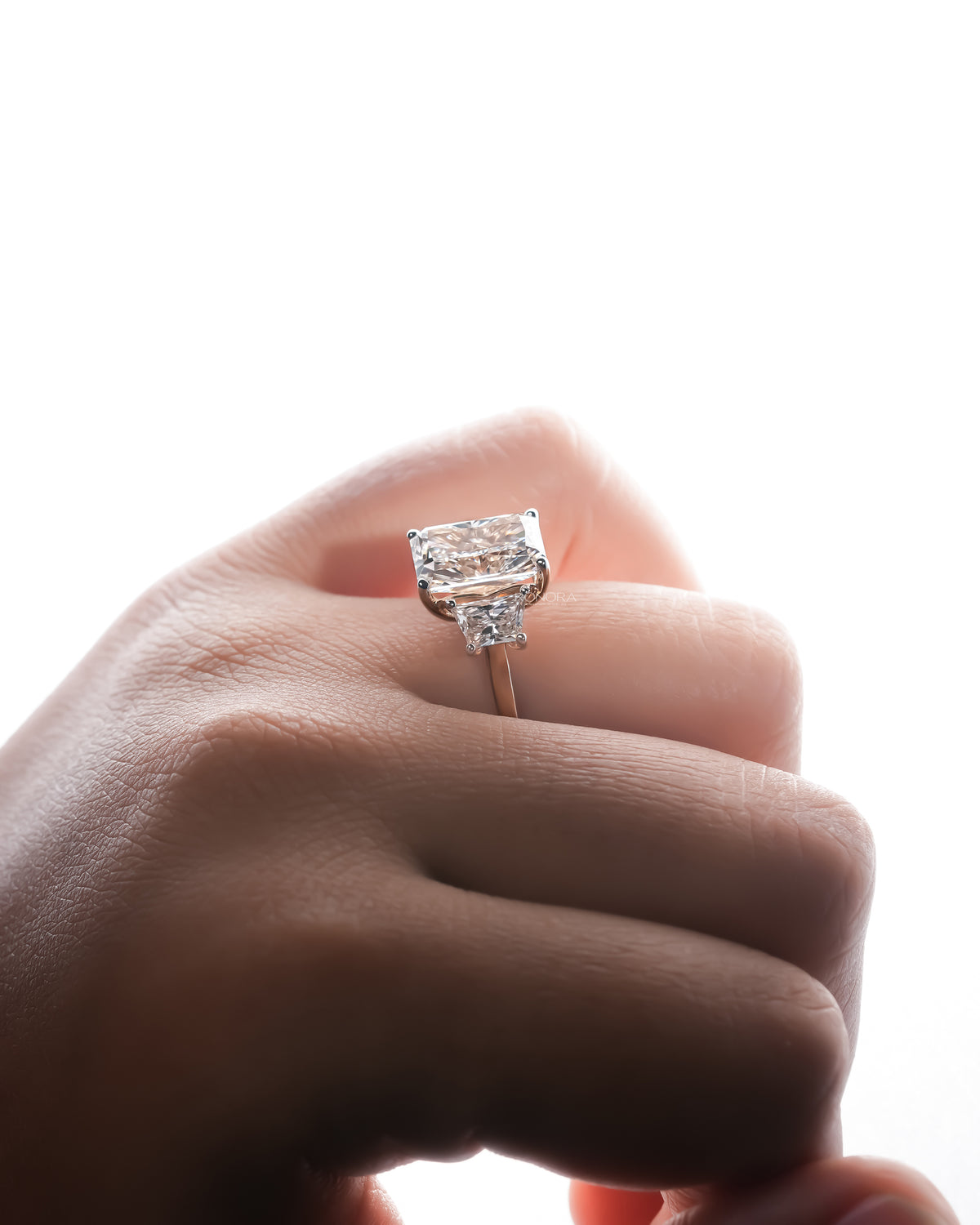 Side view of a radiant cut lab grown diamond three stone ring on a finger against a bright white background