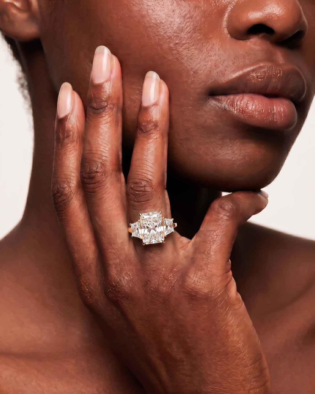 Woman with hand at her cheek wearing a radiant cut lab grown diamond three stone ring in yellow gold