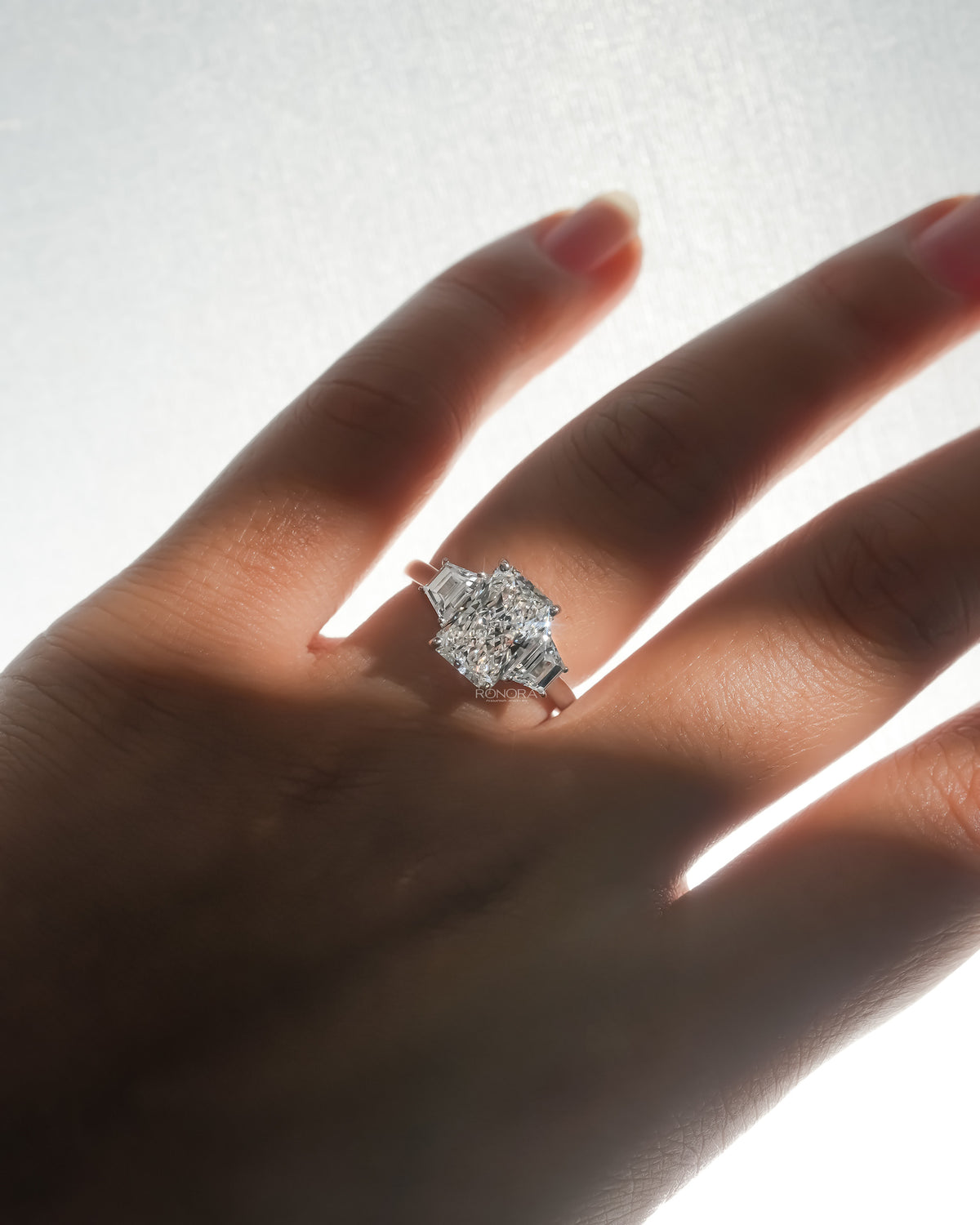 Radiant three stone lab grown diamond ring with trapezoid side stones worn on a softly backlit hand