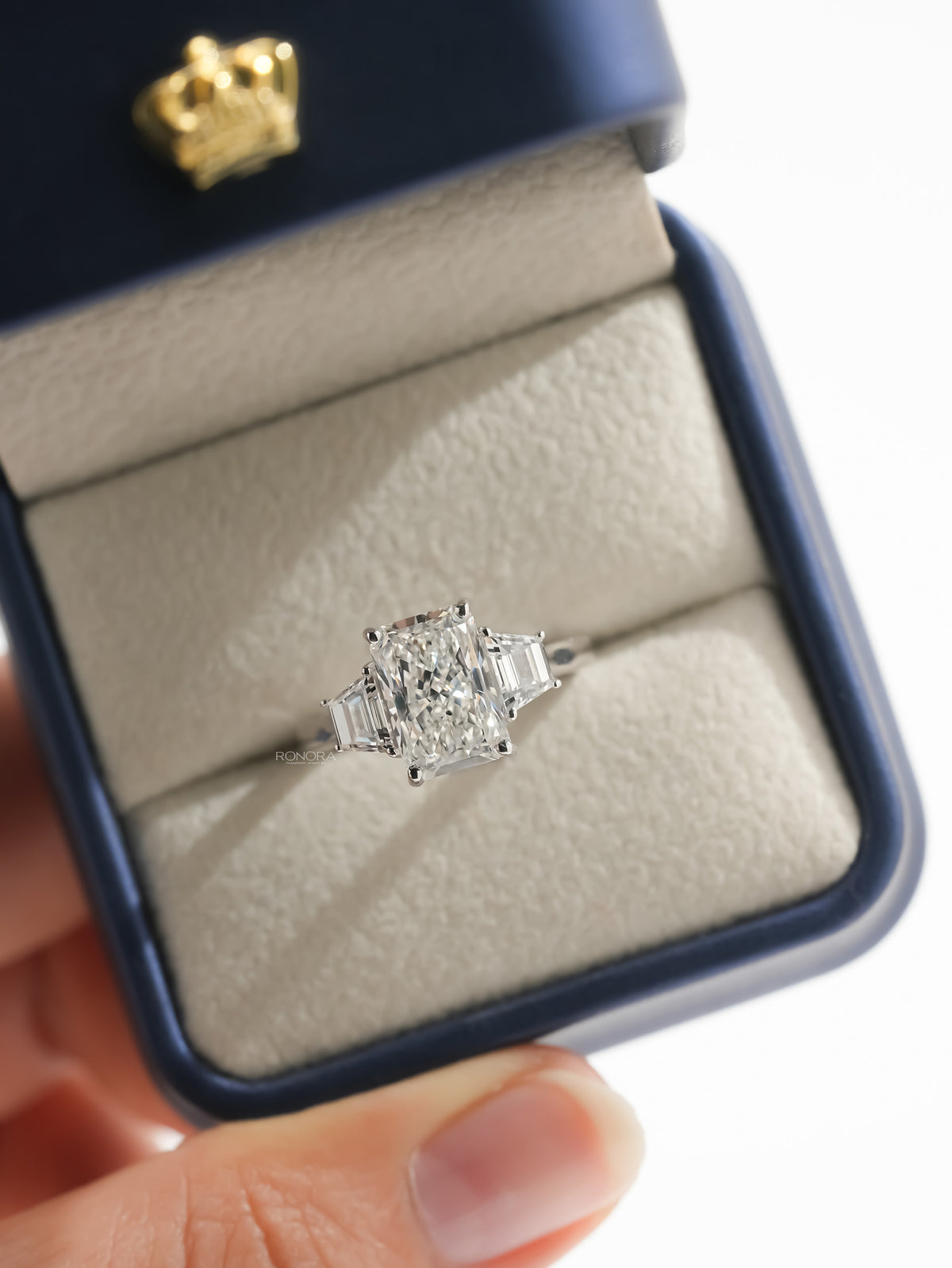 Radiant three stone lab grown diamond ring with trapezoid side stones in an open navy ring box with gold crown