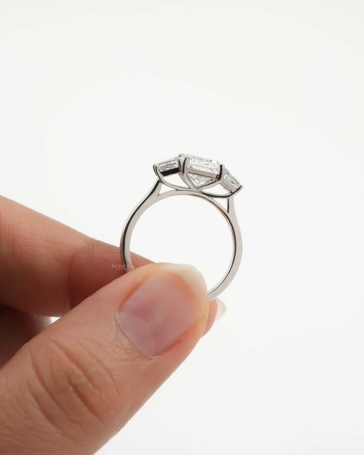 Radiant three stone lab grown diamond ring held up, showing the slim white metal band and trapezoid side stones