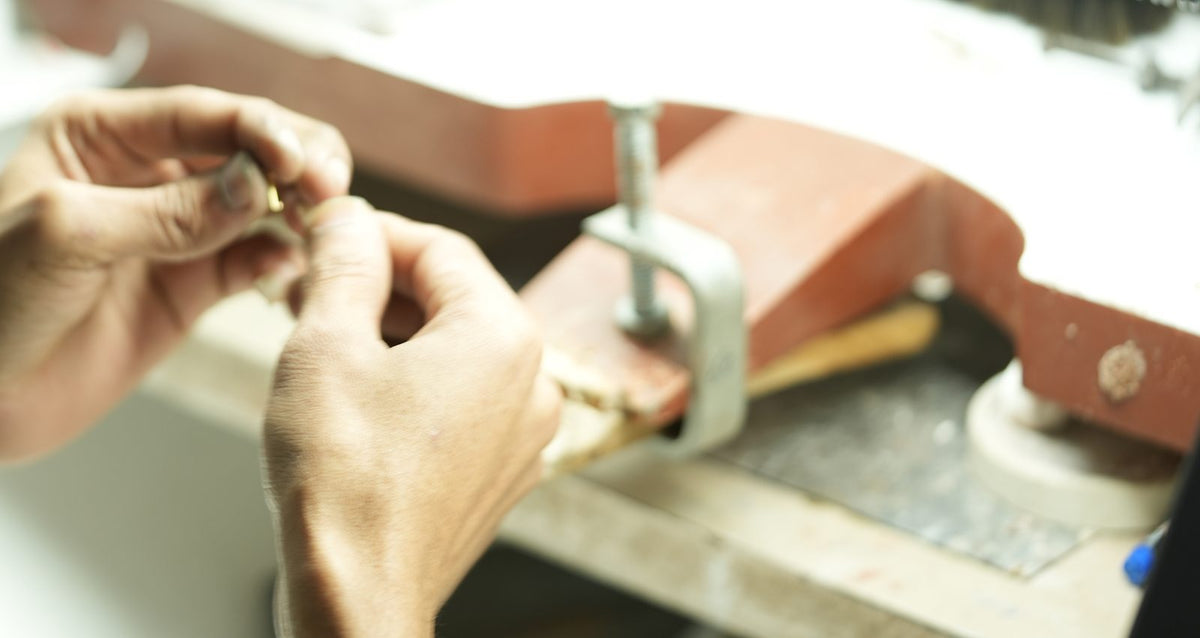 Hands at a wooden bench pin with a metal clamp, working on a small gold piece