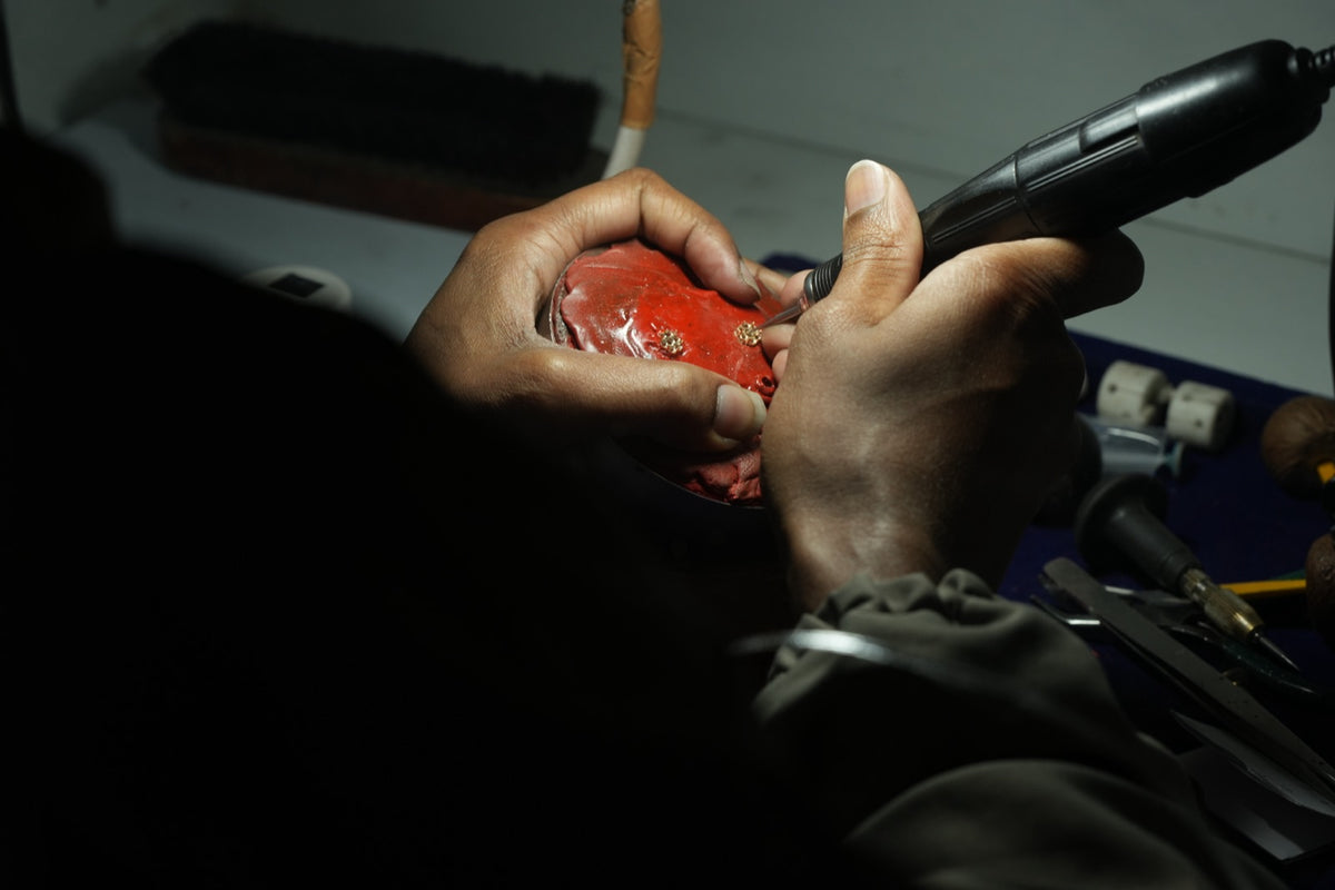 Over-the-shoulder view of a jeweler using a micro motor on small gold cluster settings held in red setting compound, tools on the bench