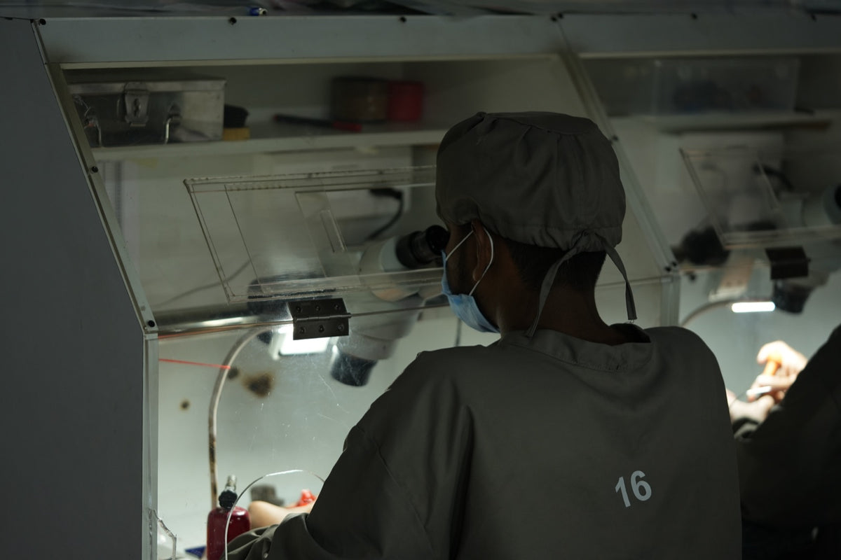 A setter in a gray uniform and cap works at a microscope station behind an acrylic shield, seen from behind