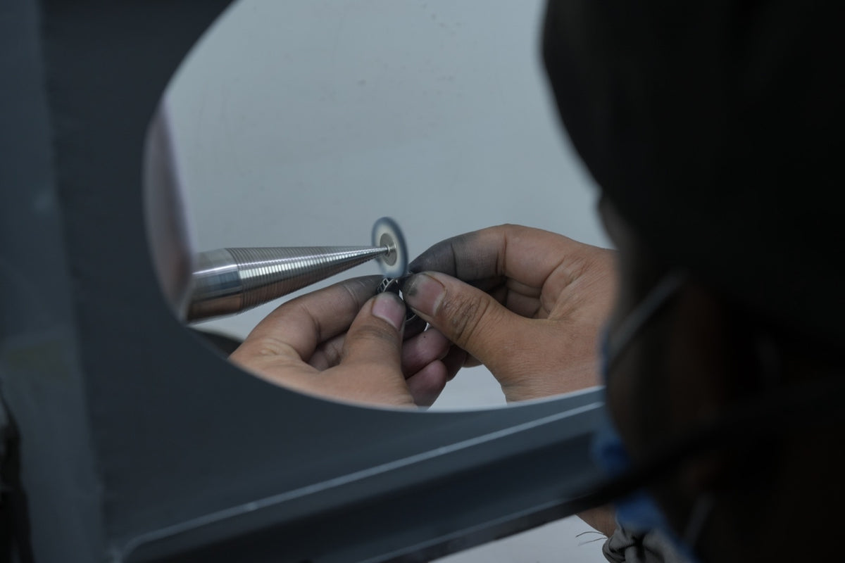View through the round window of a polishing hood: fingertips hold a ring to a small wheel on a tapered spindle
