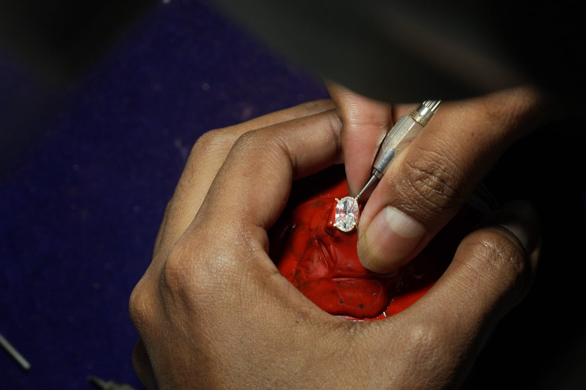 A jeweler's hands tighten a yellow gold prong around an oval stone with a hand graver, the ring held in red setting compound