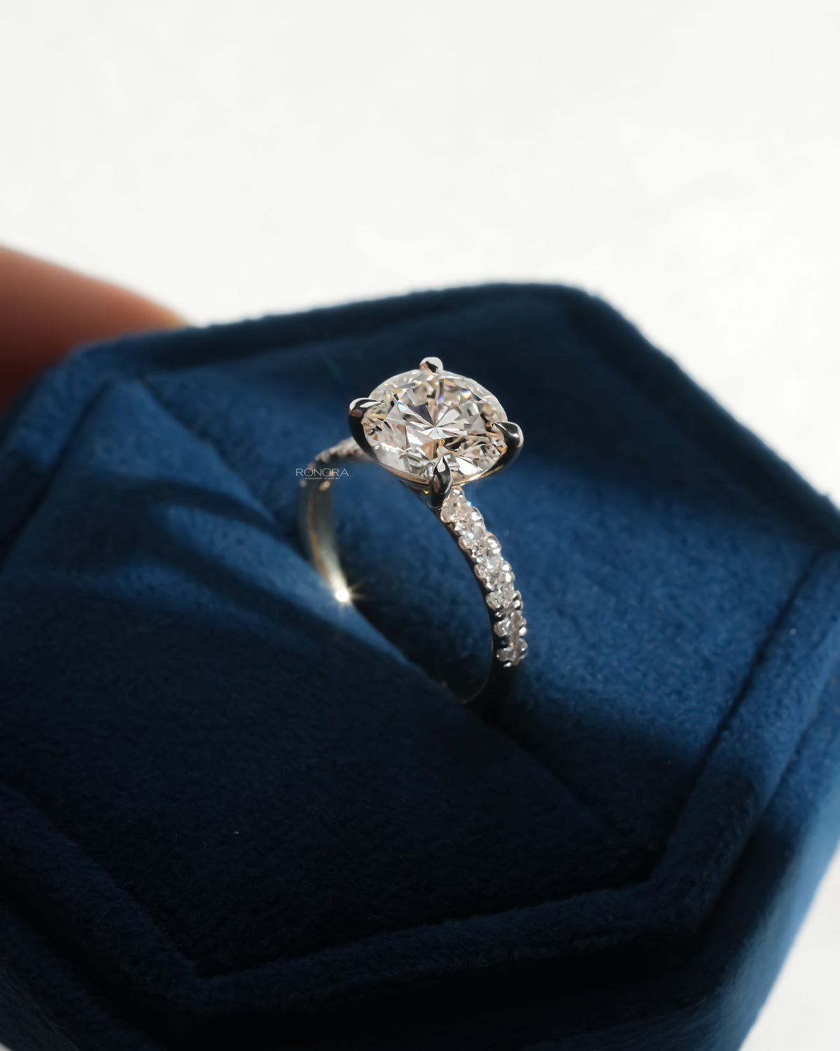 Angled view of a round hidden halo lab grown diamond ring with a pave diamond band in a blue velvet box