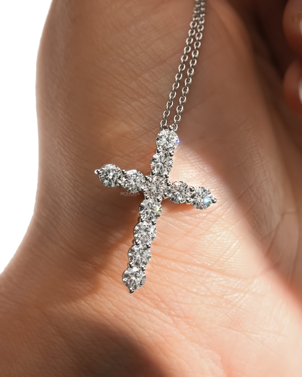 Close-up of a round lab grown diamond cross necklace with a double chain resting on skin in soft sunlight