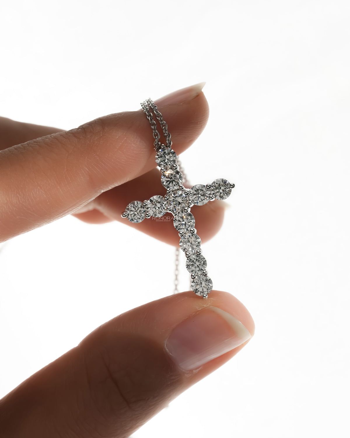 Fingers holding a round lab grown diamond cross necklace up against a white background, white metal chain