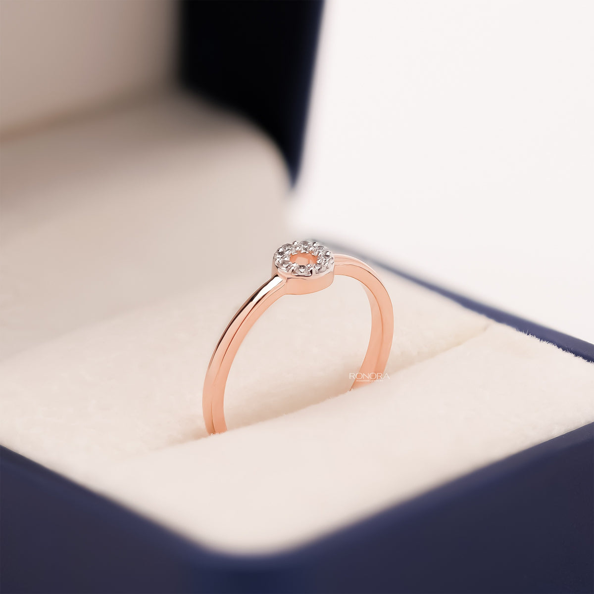 Round lab grown diamond halo ring in rose gold with a small circle of pave stones, angled in a blue and cream ring box