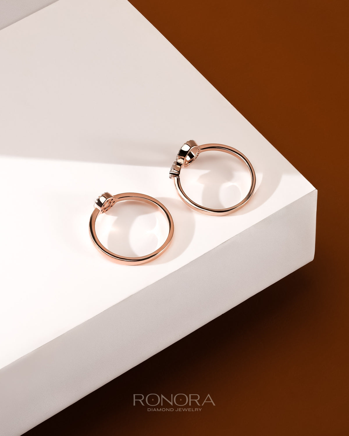 Two rose gold round lab grown diamond halo rings seen from above on a white plinth with a brown backdrop