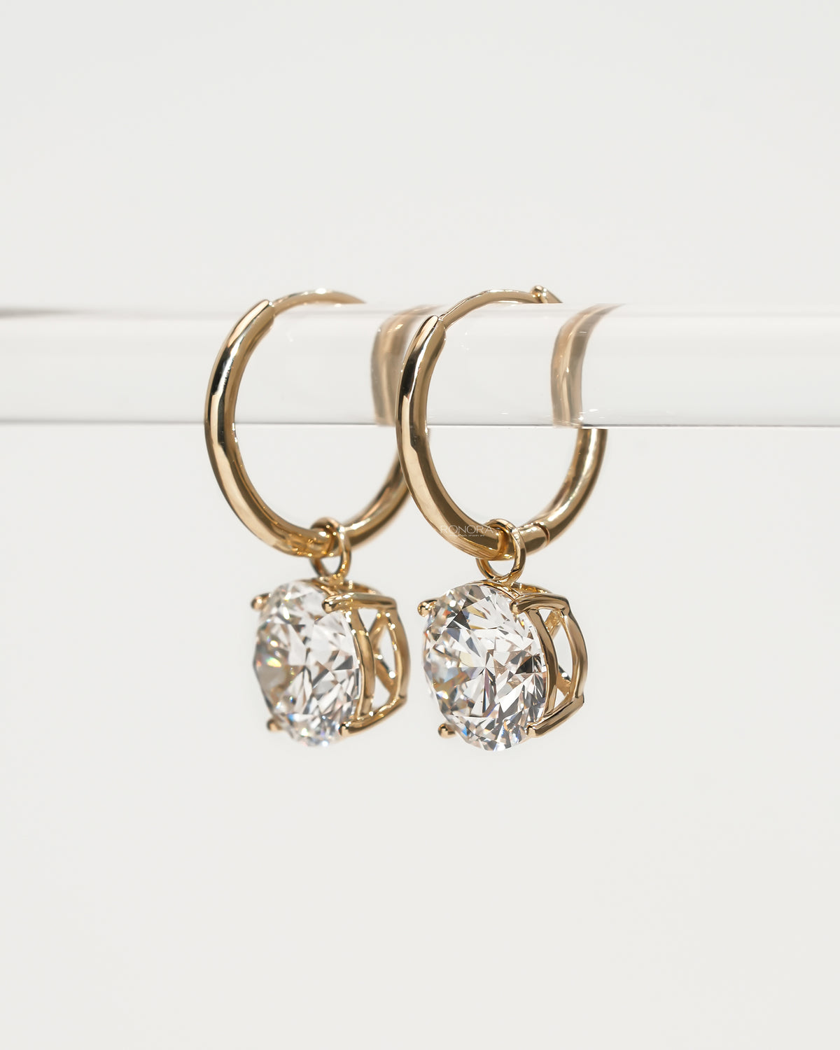 Angled view of yellow gold huggie hoops with round lab grown diamond drops hanging from a clear bar