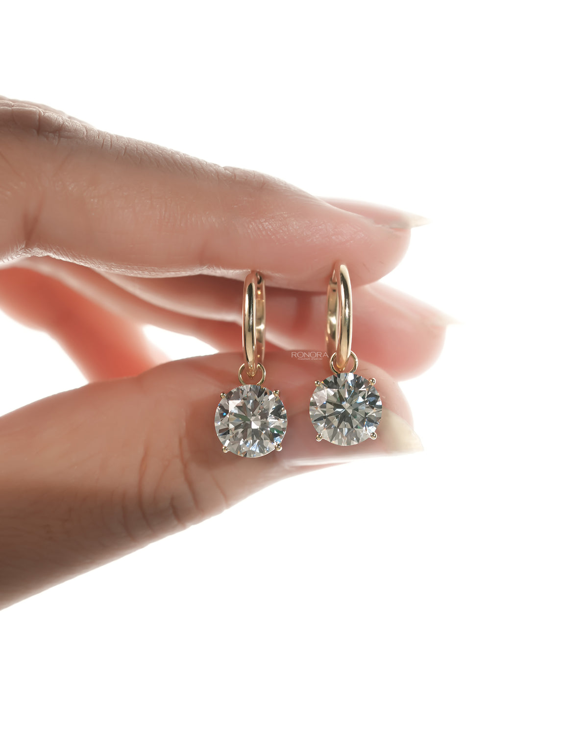 Pair of round lab grown diamond huggie drop earrings in yellow gold pinched between fingers on white
