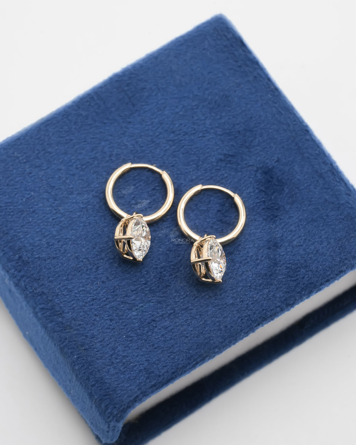 Top view of yellow gold huggie hoops with round lab grown diamond drops lying on a blue velvet box