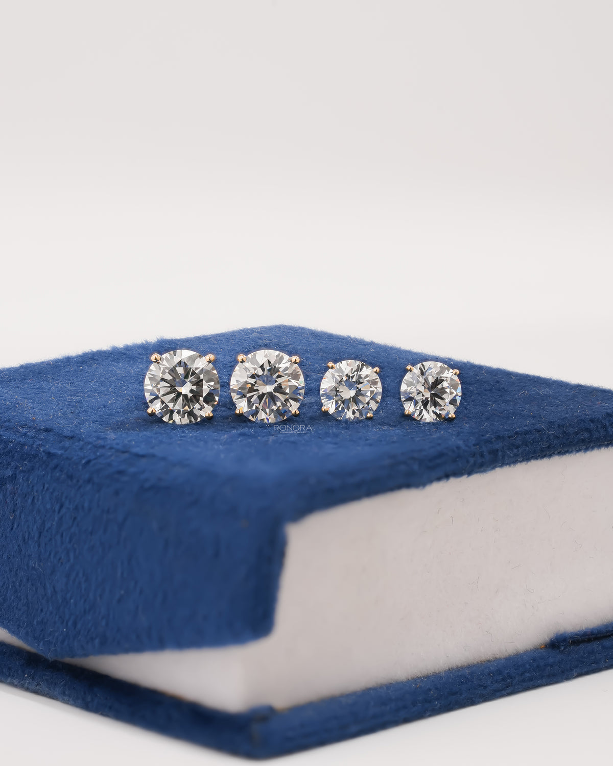 Two pairs of round lab grown diamond stud earrings in two sizes, in yellow gold four-prong settings, lined up on a navy velvet box