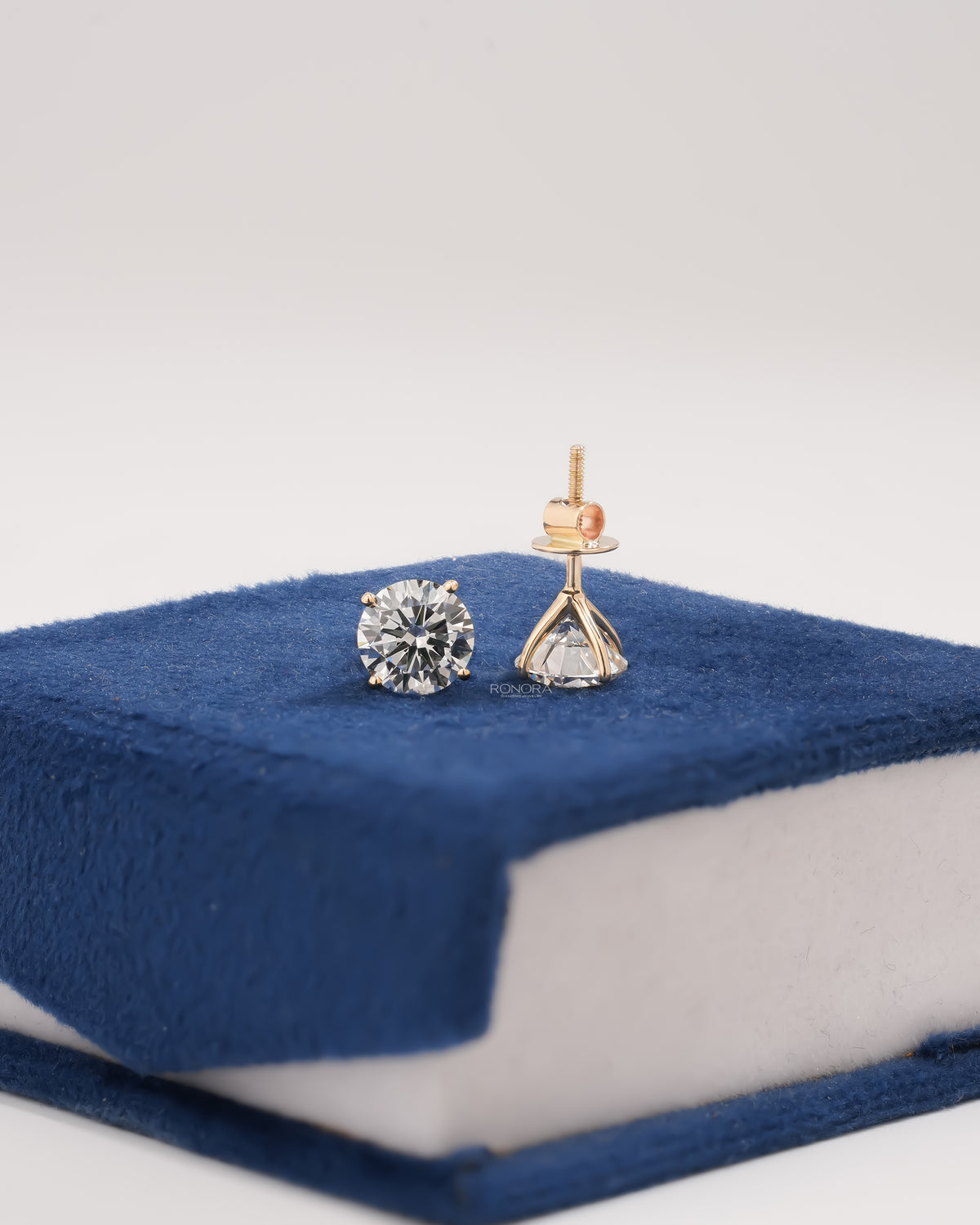 Round lab grown diamond stud earrings, one showing the front and one the screw back, on blue velvet
