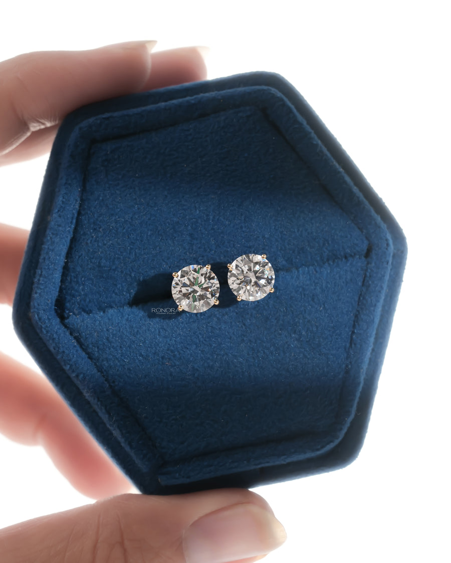 Round lab grown diamond stud earrings in four prong yellow gold shown in a hexagonal blue velvet jewelry box