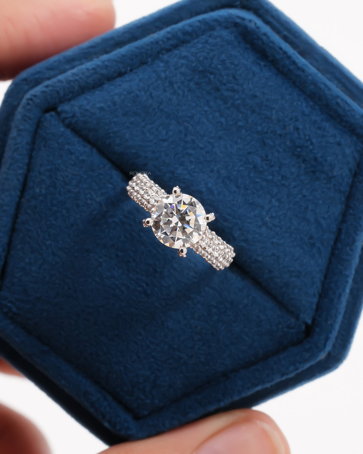 White gold ring with a round moissanite in white prongs on a three-row pave band, in a navy velvet box
