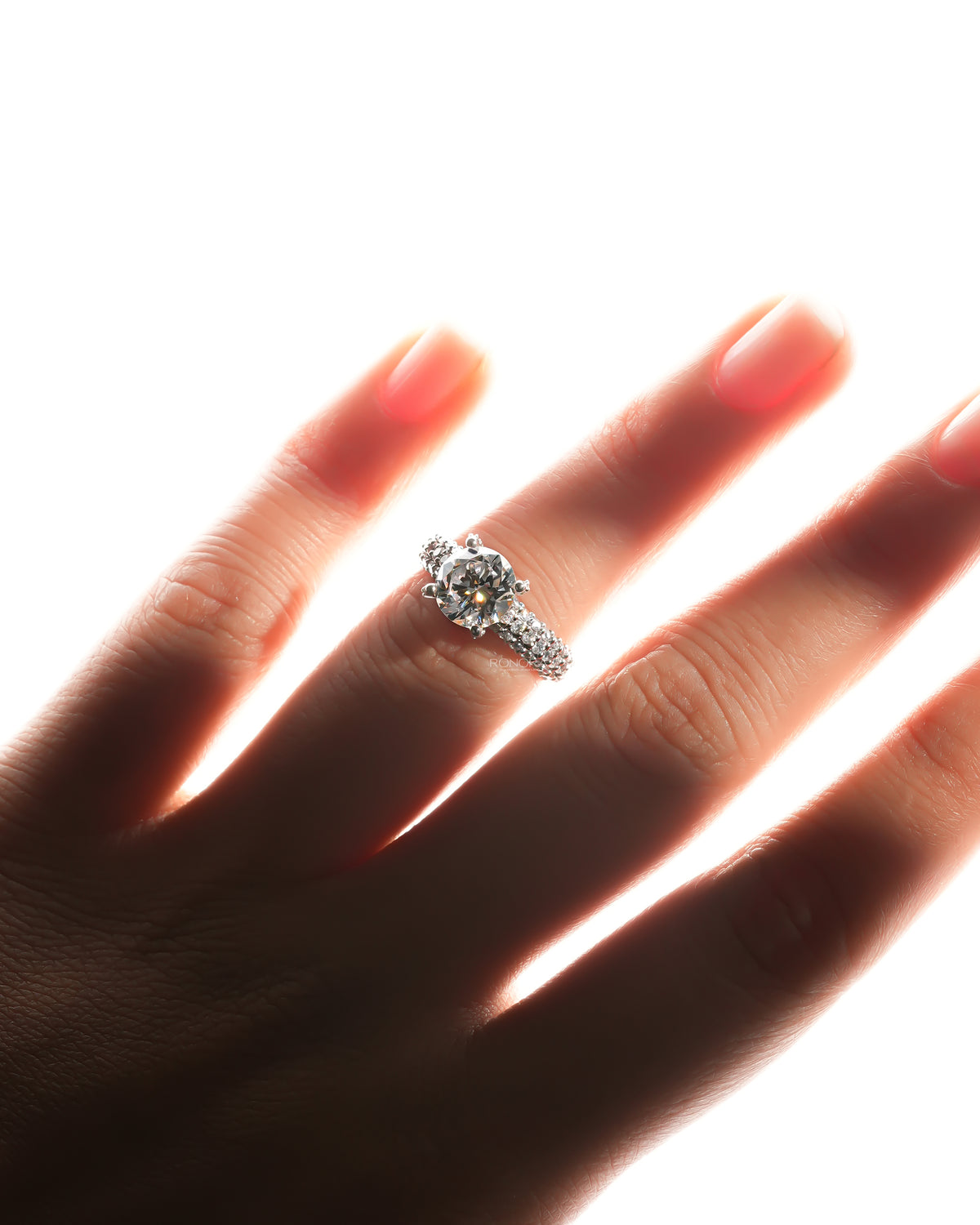 Hand wearing a round moissanite engagement ring with a pave band, backlit and glowing against a bright background