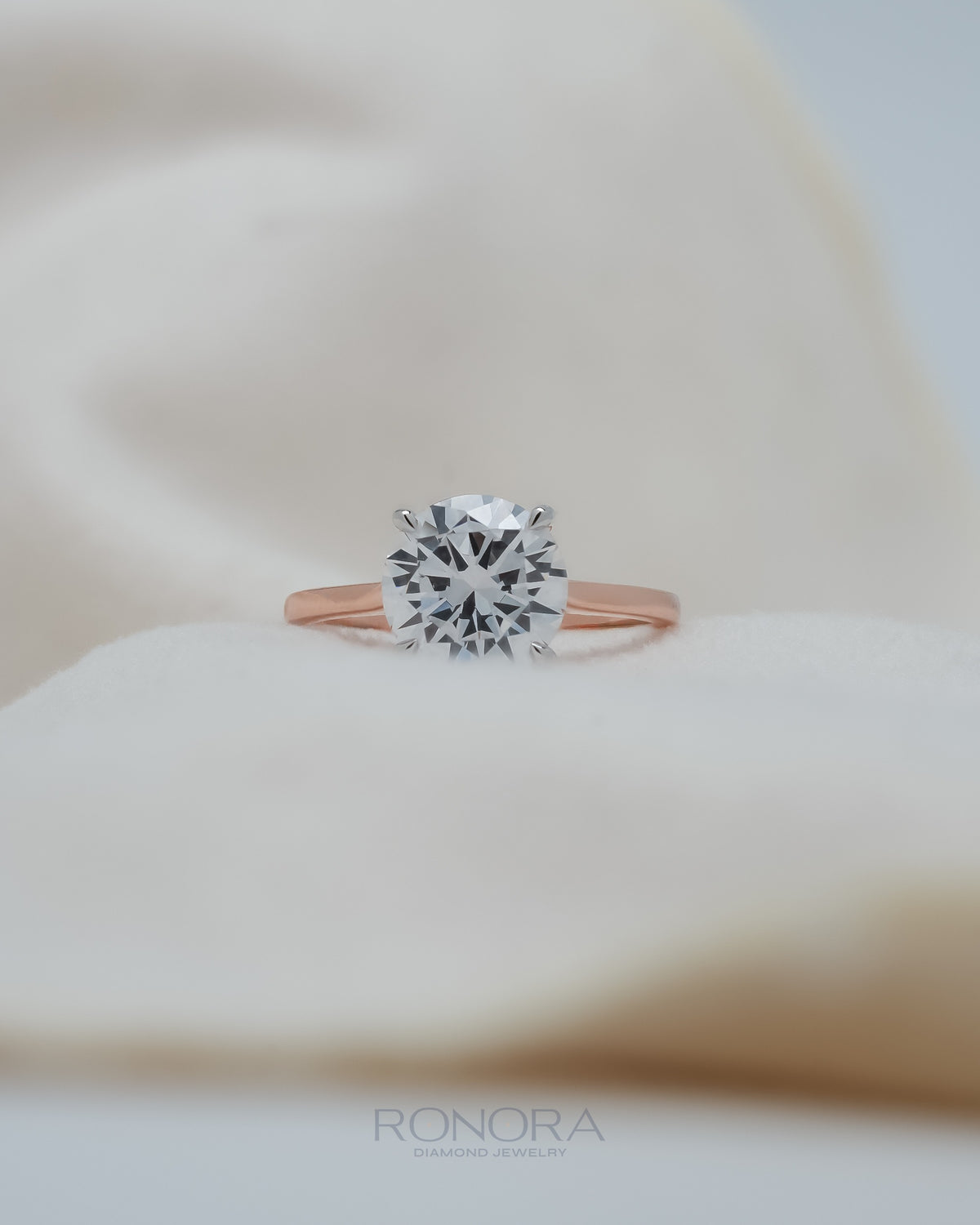 Round moissanite solitaire ring with a large four prong stone on a rose gold band, resting on soft cream fabric
