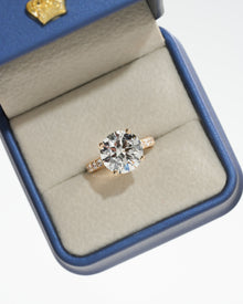 Round pave lab grown diamond engagement ring with a gold pave band in a blue ring box, top view