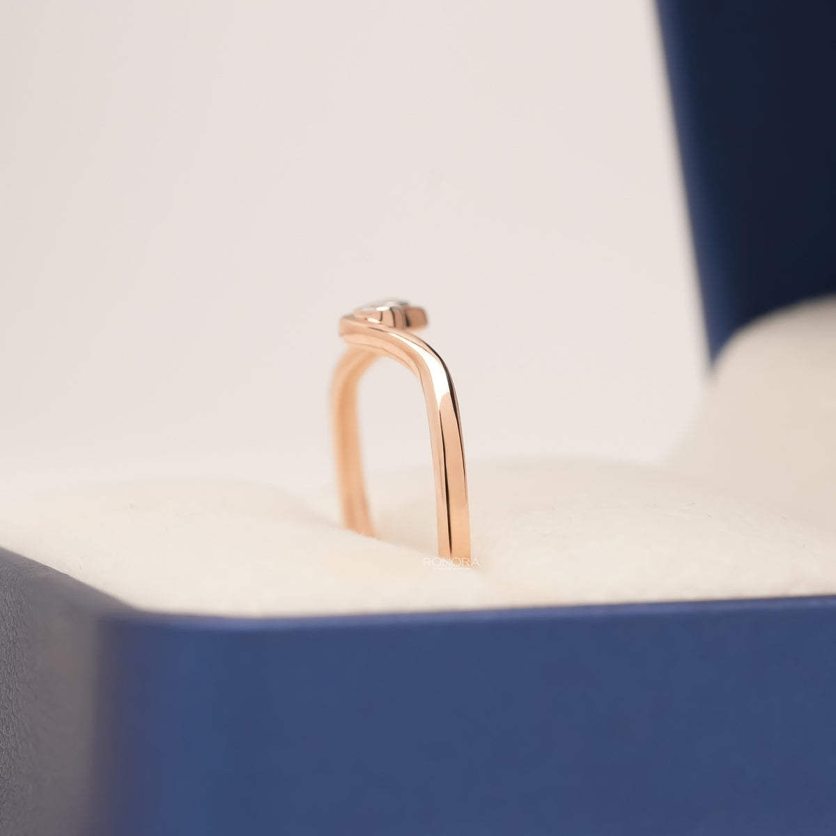 Side view of a rose gold two stone bezel lab grown diamond ring with a wave design standing in a blue ring box