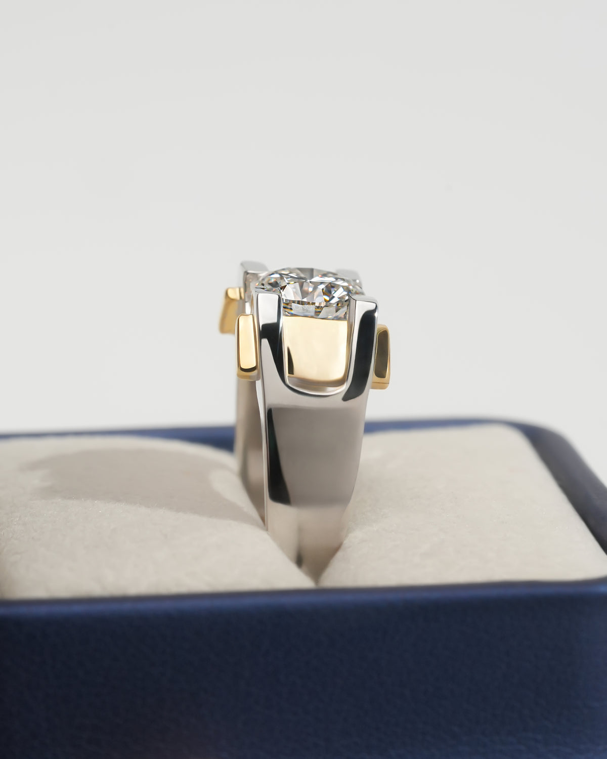 Side view of a round lab grown diamond solitaire engagement ring with a wide two tone gold band in a navy ring box
