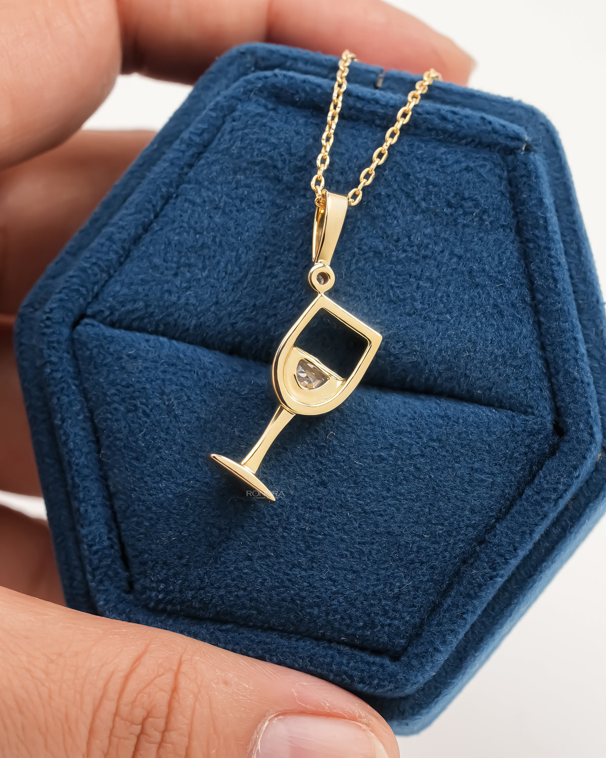 Back view of a yellow gold wine glass lab grown diamond pendant on a blue velvet box held in a hand