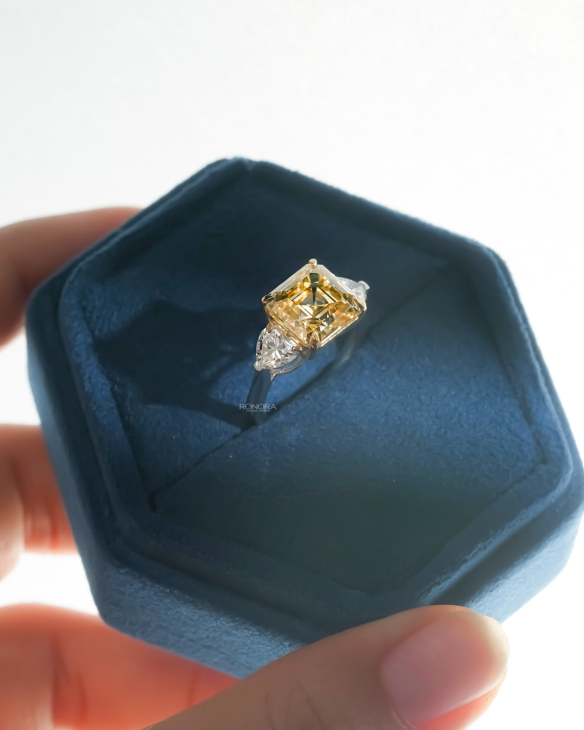 Asscher yellow lab grown diamond ring with heart side stones angled in a blue velvet hexagon ring box
