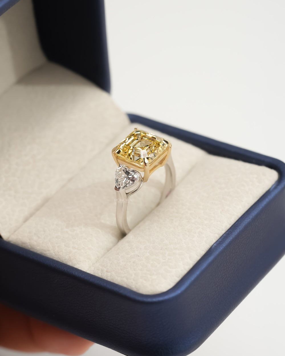 Asscher yellow lab grown diamond ring in an open navy ring box, yellow gold prongs and a white metal band