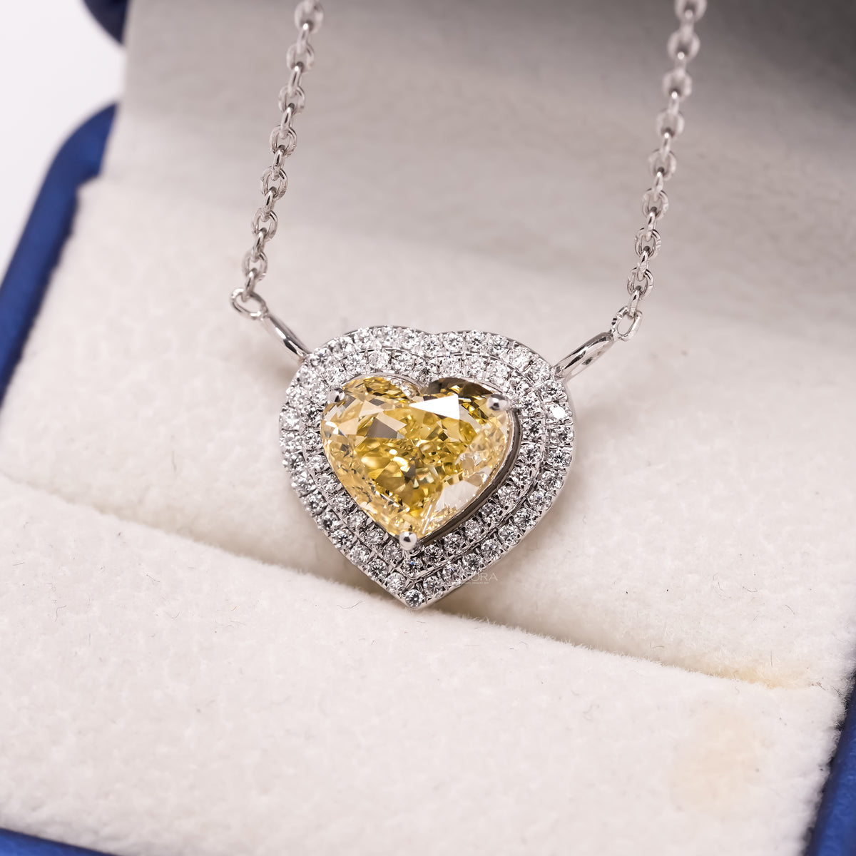 Close-up of a yellow heart cut lab grown diamond necklace with a double halo in a cream and blue jewelry box