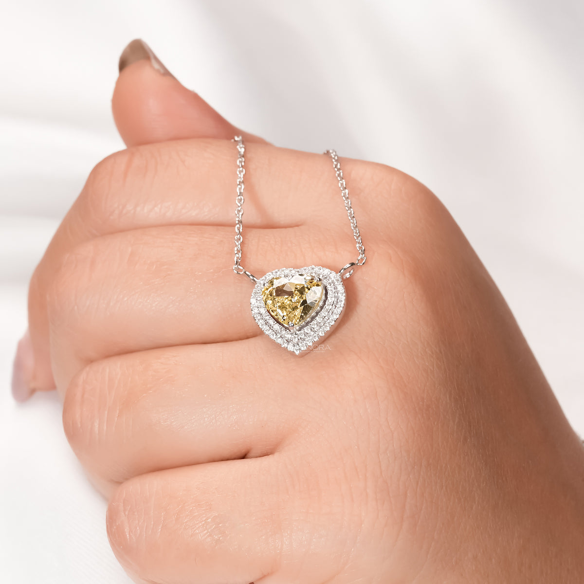Hand wearing a yellow heart cut lab grown diamond necklace with a double halo pendant resting on the knuckles