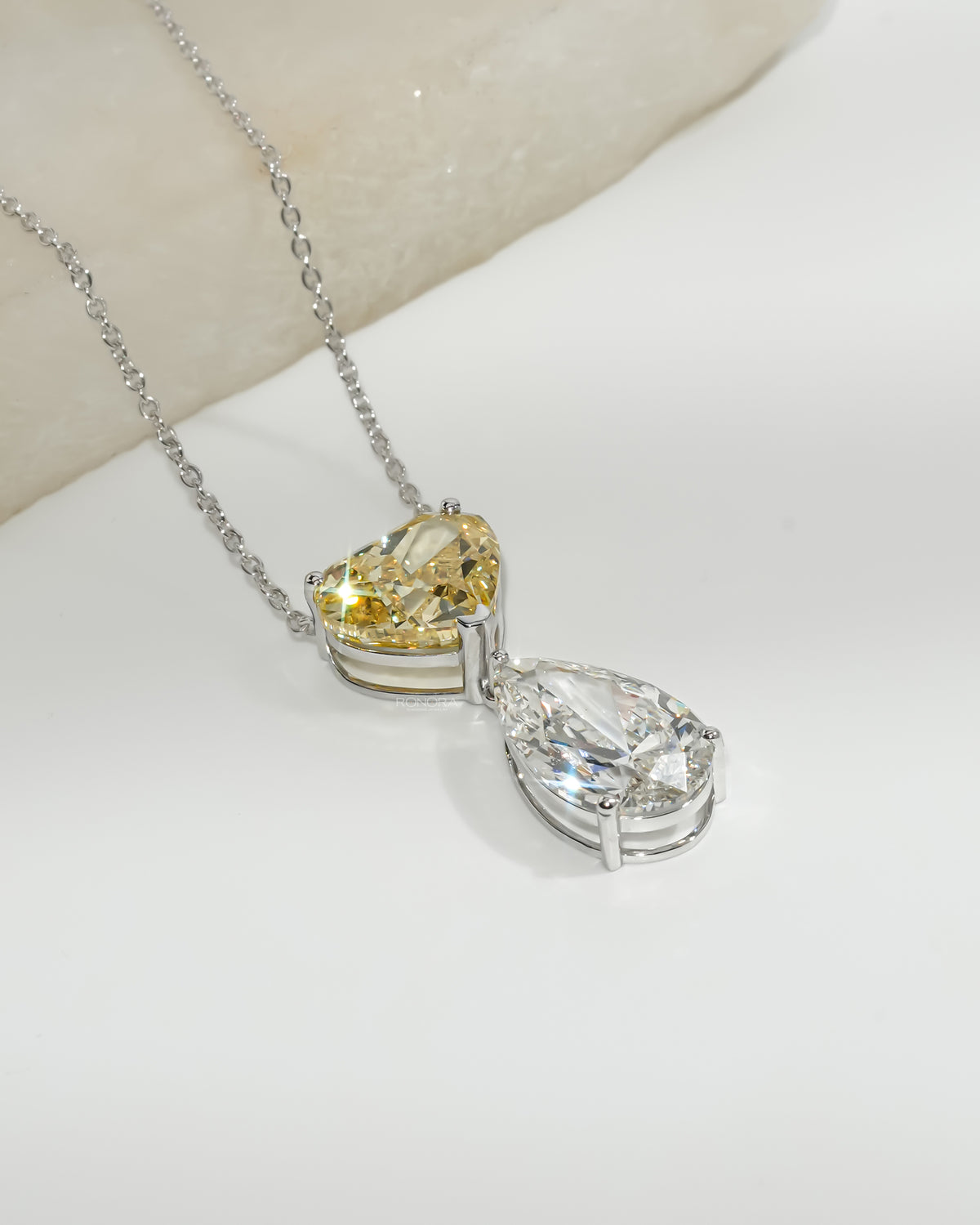 Close-up of a yellow heart and white pear lab grown diamond necklace on a white gold chain, resting on a white stone