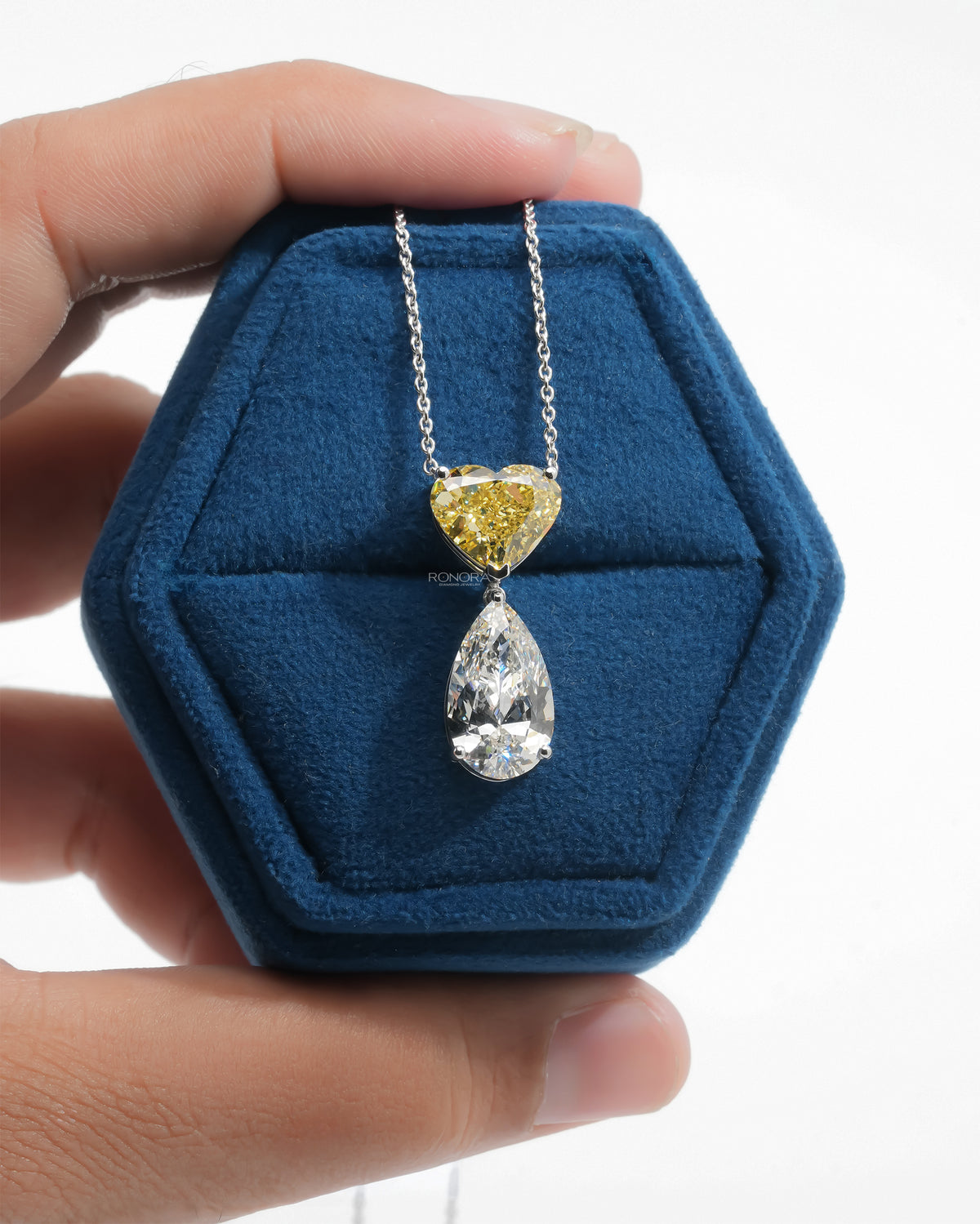 Hand holding a blue velvet jewelry box with a yellow heart and white pear lab grown diamond necklace in white gold