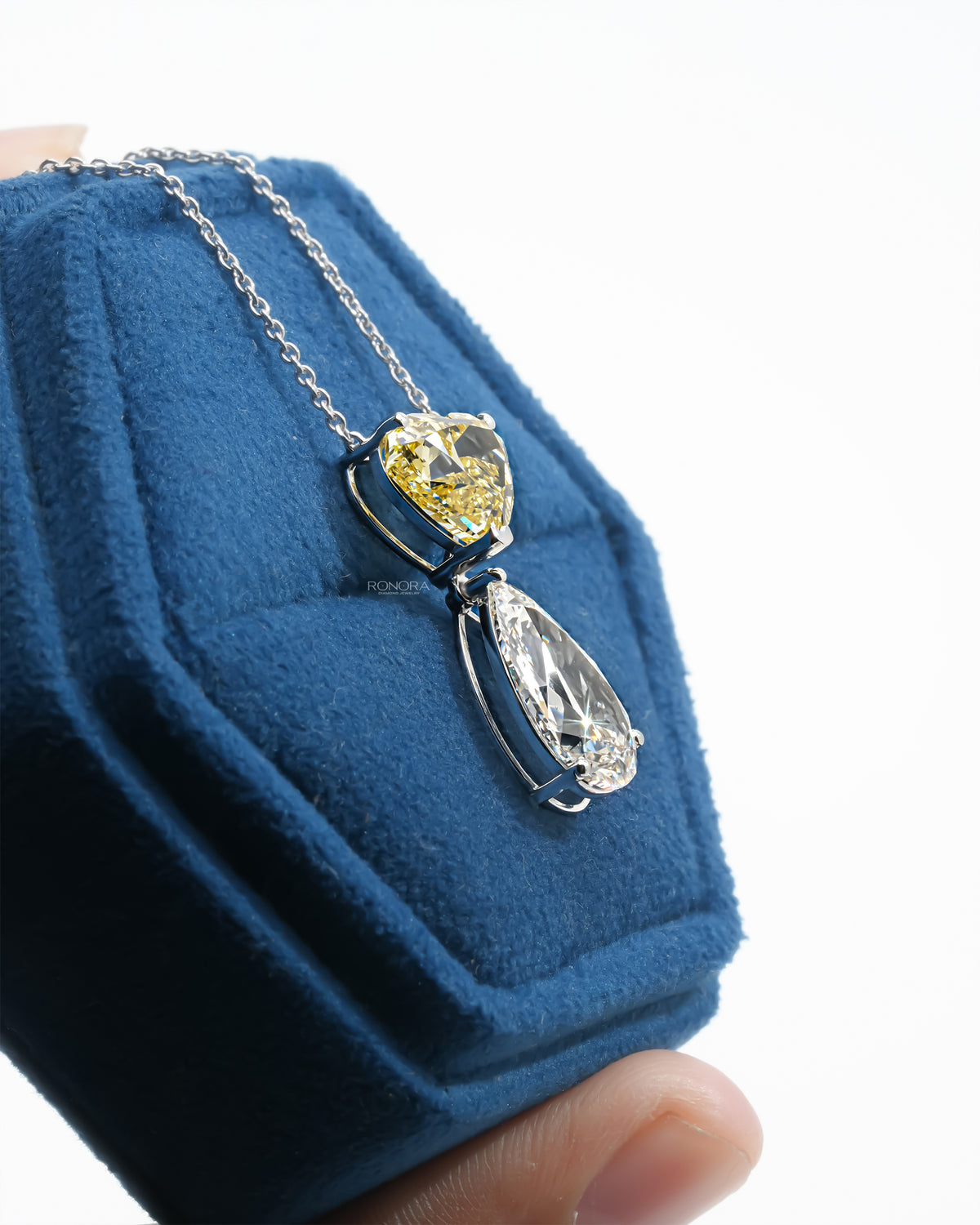 Side view of a yellow heart lab grown diamond necklace with a white pear drop, set on a blue velvet jewelry box
