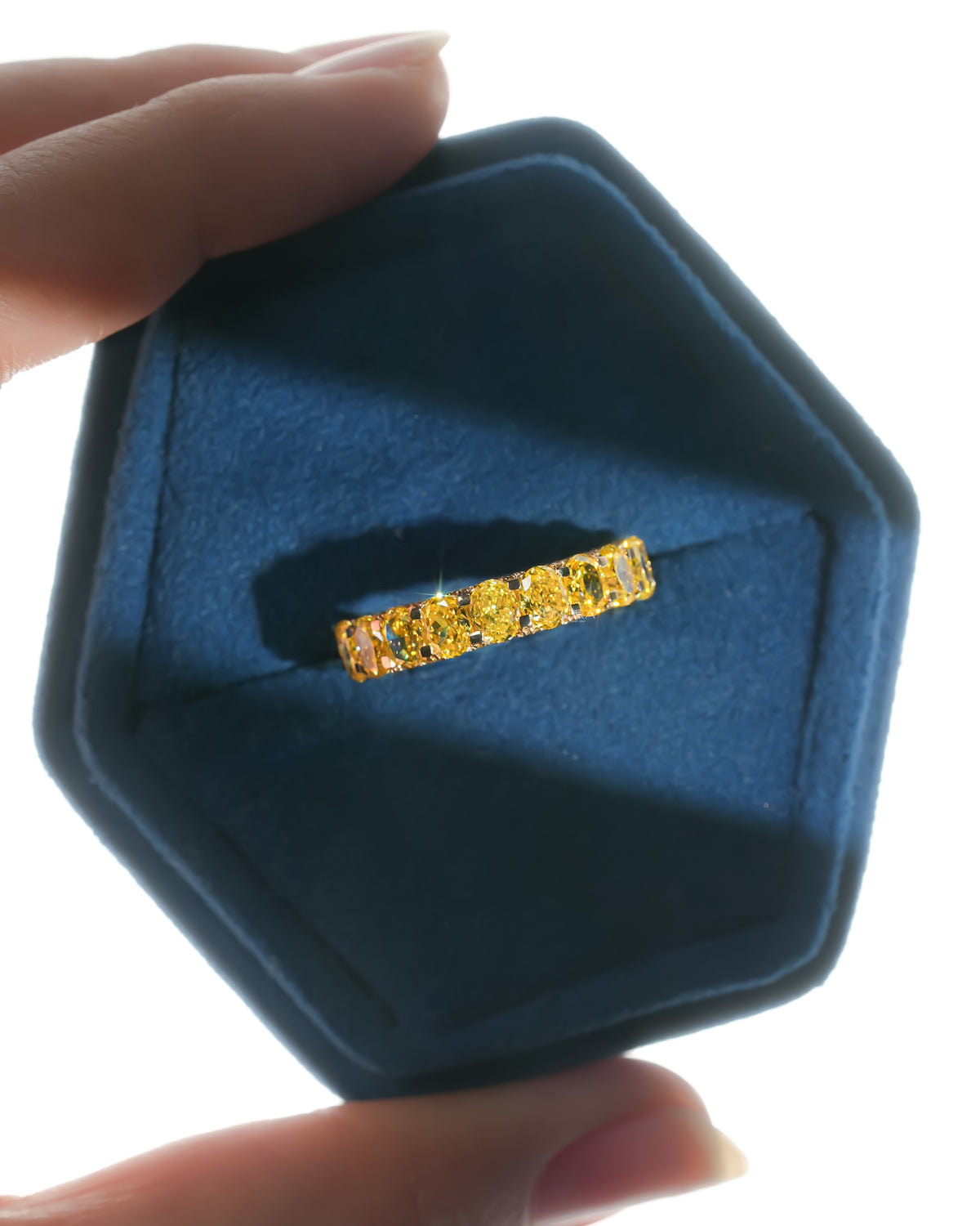 Yellow lab grown diamond oval eternity band in a blue velvet hexagon ring box held between two fingers, top view