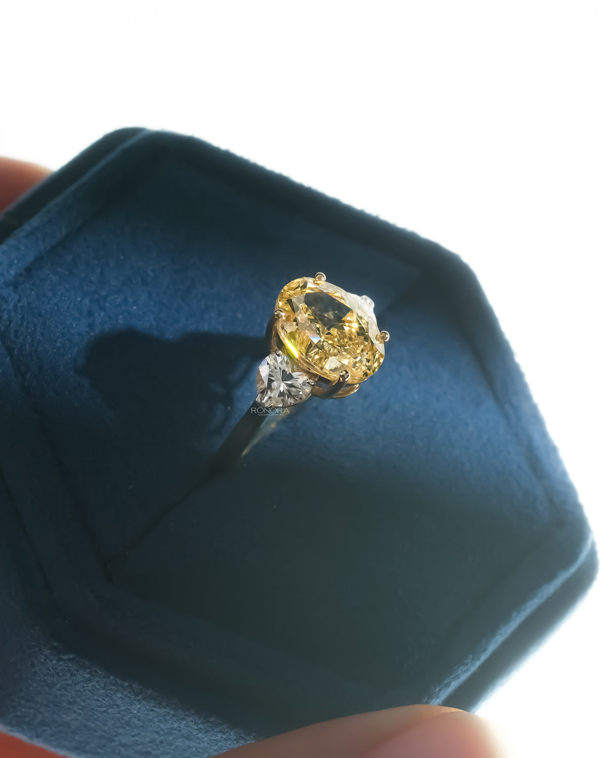 Oval yellow lab grown diamond ring with heart side stones angled in a blue velvet hexagon jewelry box
