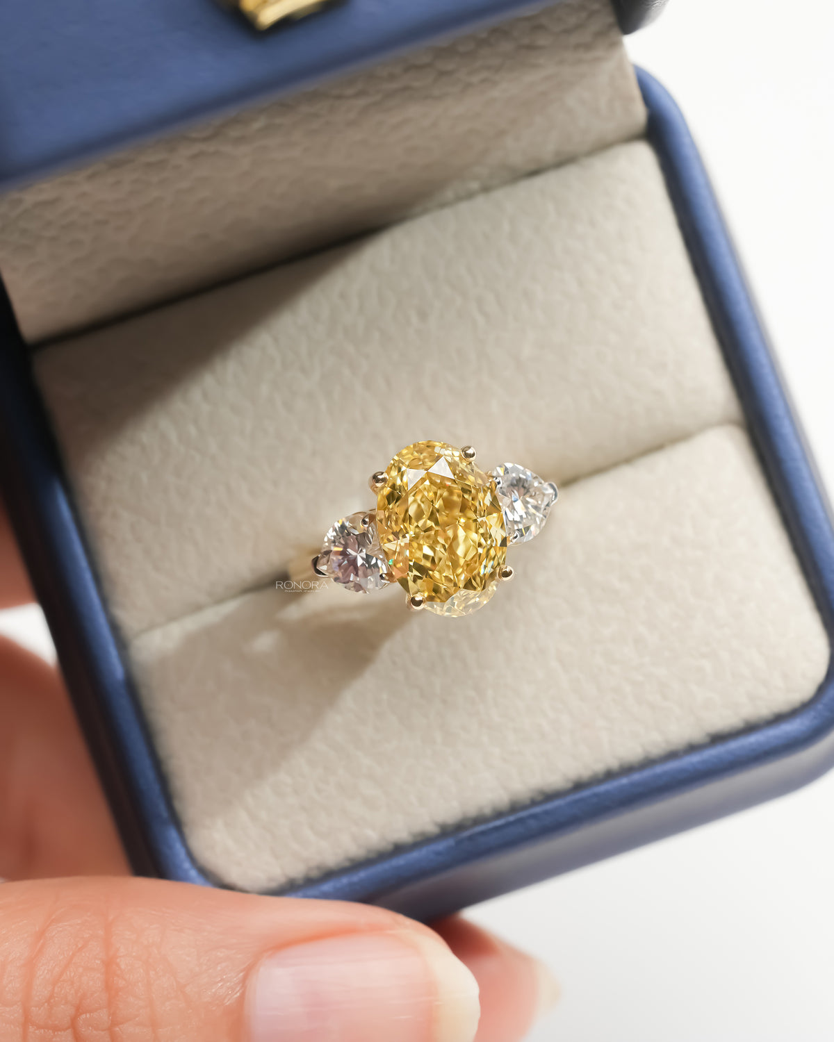 Oval yellow lab grown diamond ring with two heart side stones in an open navy ring box with cream cushion