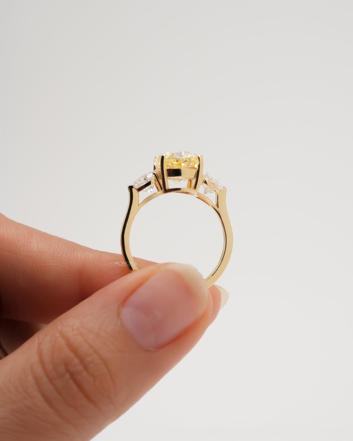 Side profile of a yellow gold oval lab grown diamond ring held between fingertips, showing band and side stones